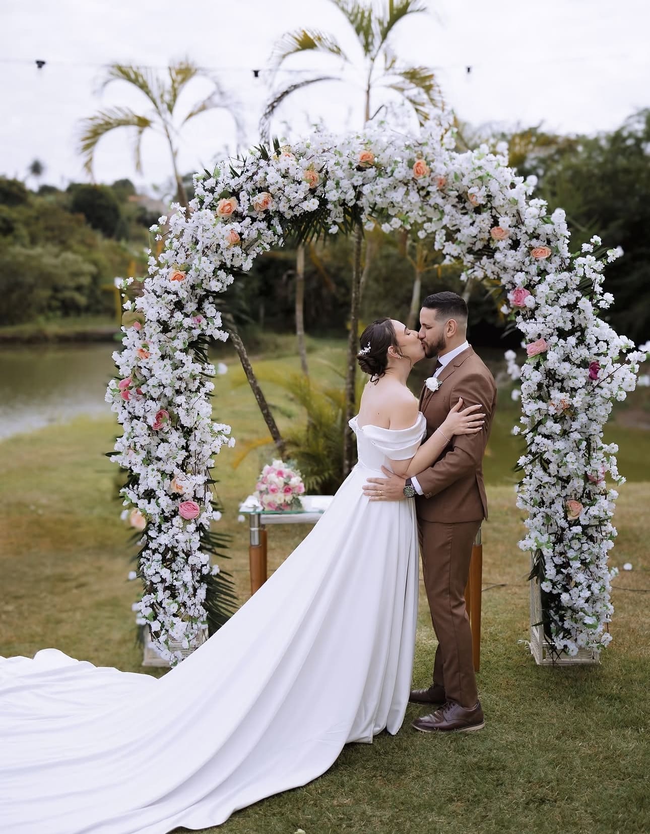 Casal se beijando sob arco floral à beira da lagoa no Laurus Eventos