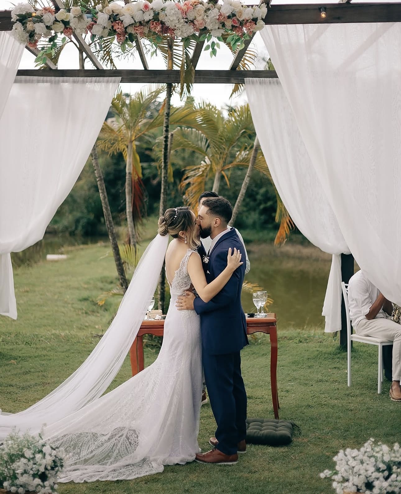 Noivos se beijando sob pérgola com cortinas brancas à beira da lagoa