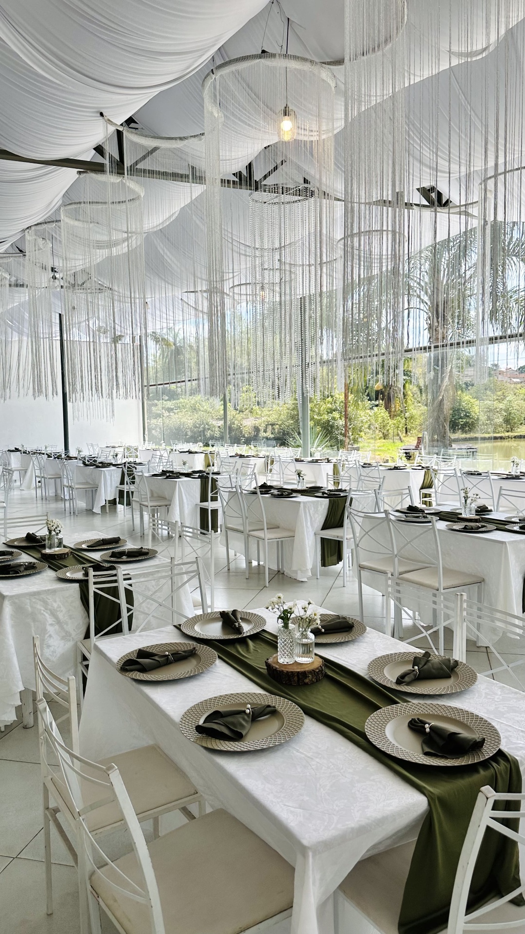 Vista ampla do salão com cortinas de cristal, mesas em verde-oliva e janelas para o jardim