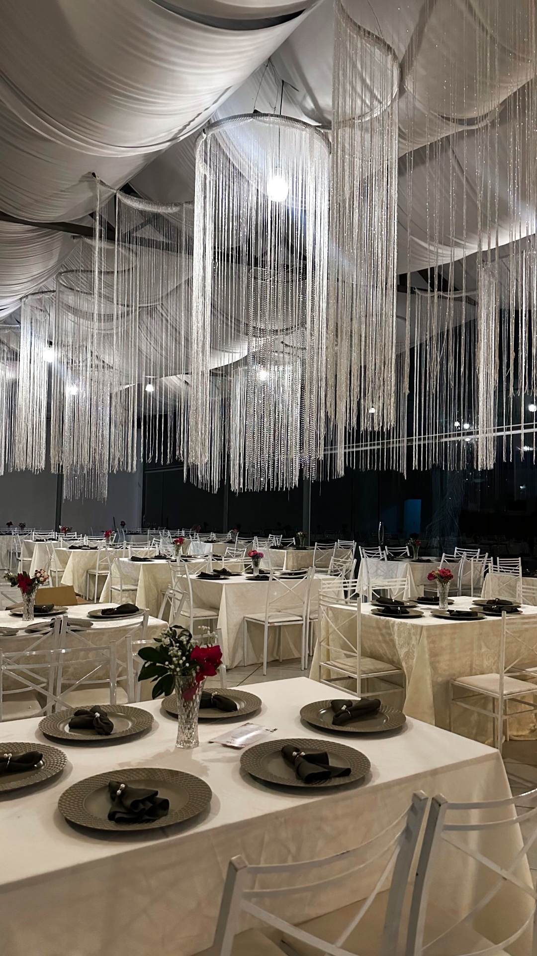 Salão à noite com lustres de franjas brilhantes e mesas decoradas em preto e flores